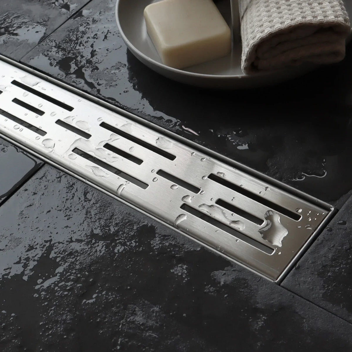 Stainless steel linear shower drain on wet dark tile floor with soap and towel nearby
