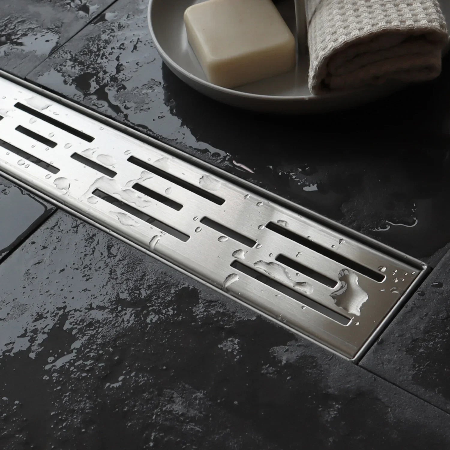 Stainless steel linear shower drain on wet dark tile floor with soap and towel nearby