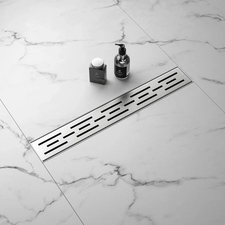 Stainless steel linear shower drain on marble tile bathroom floor with soap and dispenser.
